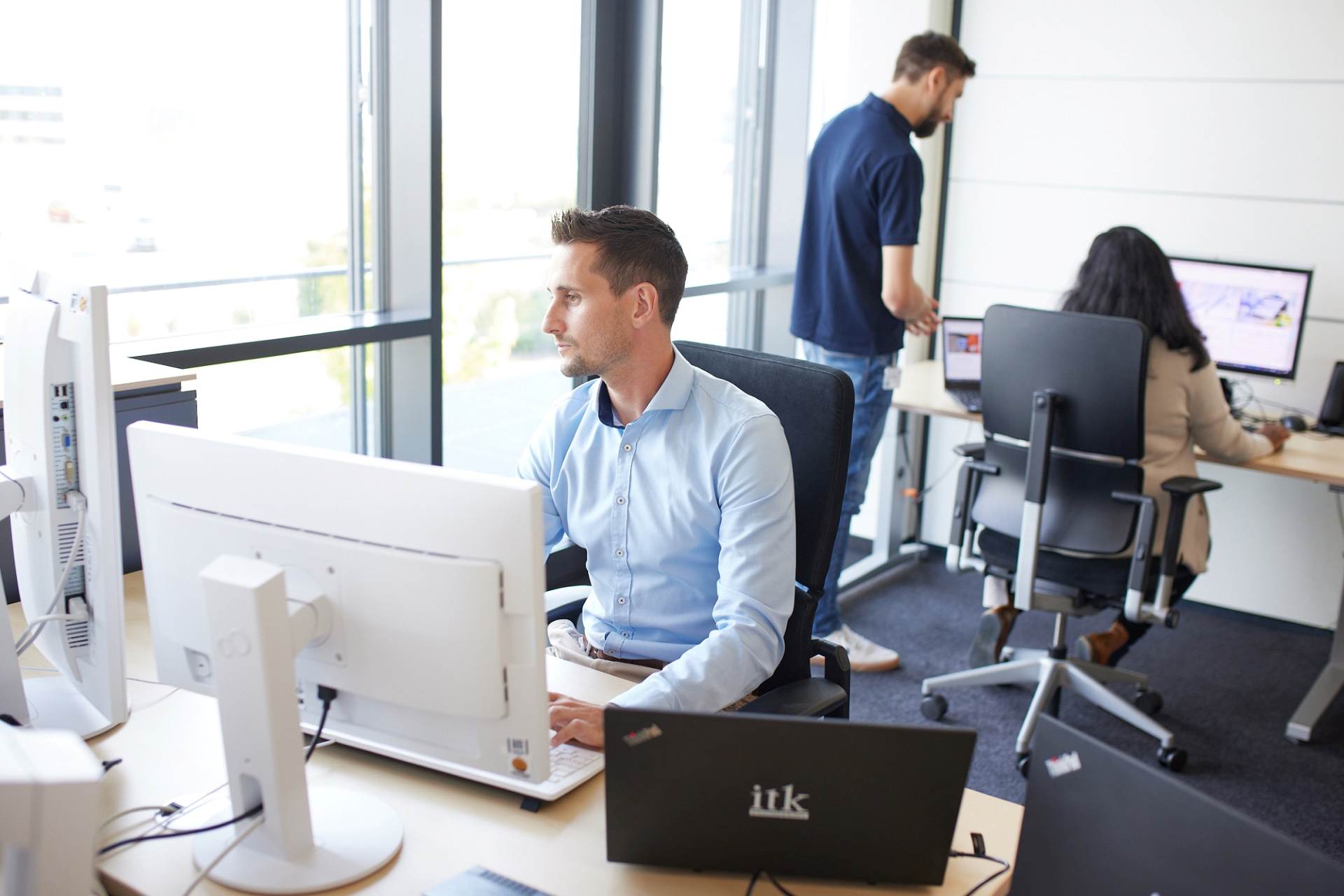 An engineer is working on a laptop; two colleagues are working in the background