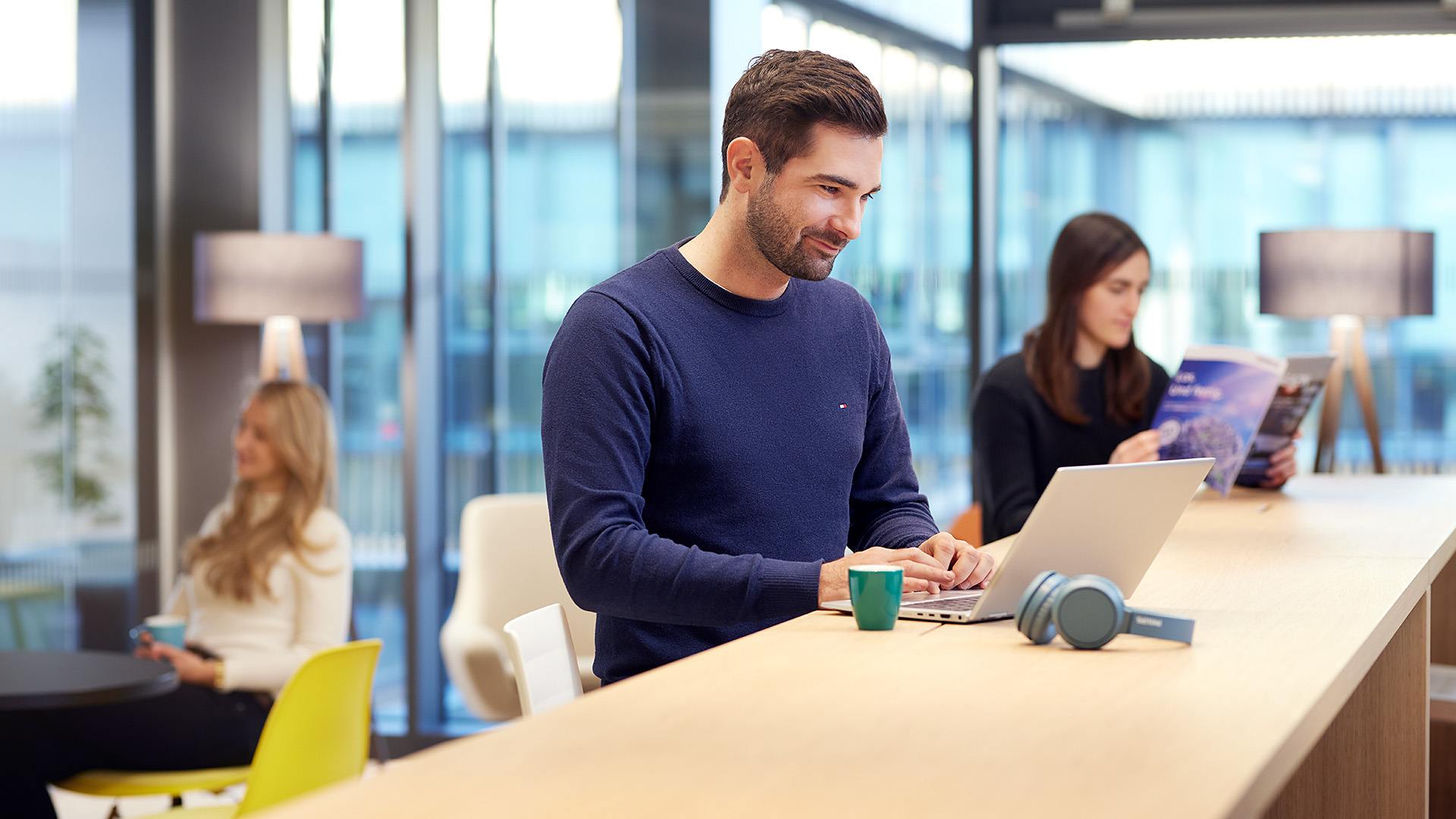 ITK Mitarbeiter arbeitet am Laptop in der Kaffeeküche. 2 ITK Mitarbeiterinnen sitzen im Hintergrund.