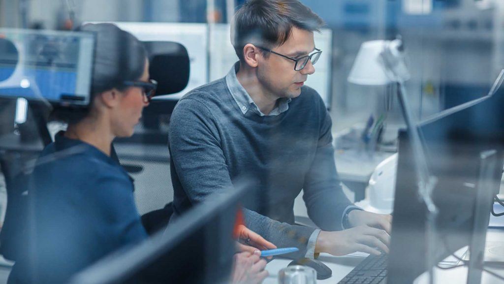Aufnahme durch Glas in einer modernen Fabrik: Weiblicher Projektleiter im Gespräch mit einem männlichen Wirtschaftsingenieur, der an einem Personal Computer arbeitet.