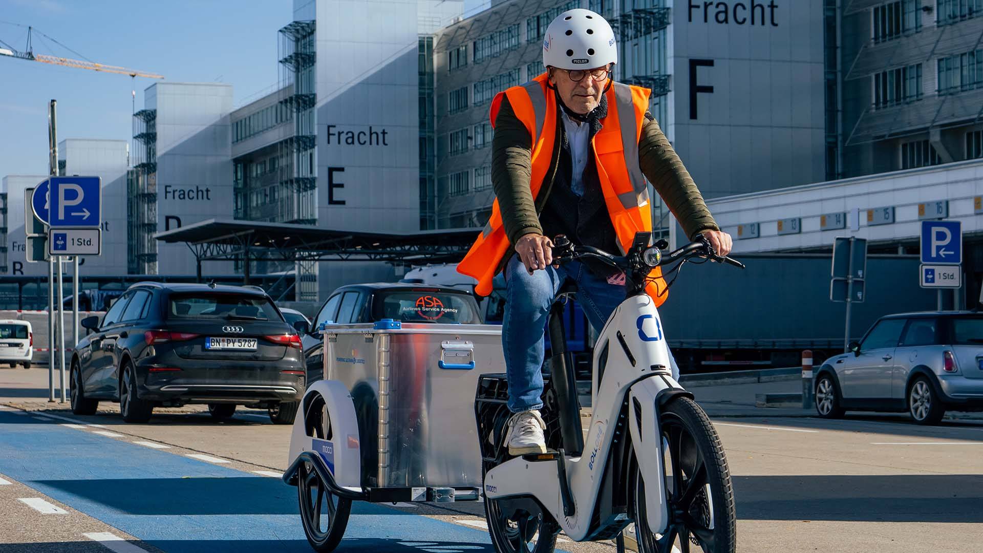 A man is cycling on the e-bike mocci across an airport area.