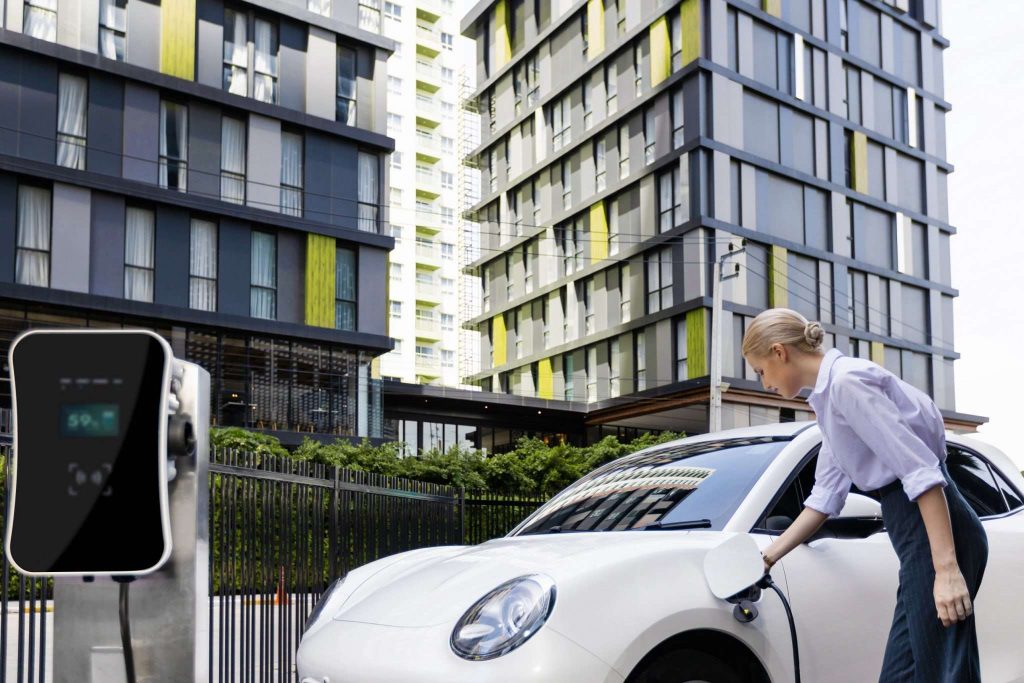 Progressive businesswoman inserts charger plug from charging station to her electric vehicle with apartment condo building in background. 