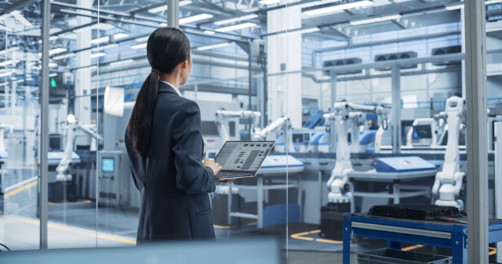 Eine Frau im Business-Outfit überwacht mit einem Laptop Roboterarme an einer Fertigungslinie – als ganzheitlicher Entwicklungspartner gestaltet ITK eingebettete Systeme als standardisierte und integrierte, zentrale Schnittstellen.
