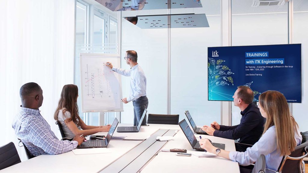 Participants in an ITK training sit at a table in a meeting room while the trainer explains something on a flipchart. A screen can be seen in the background.