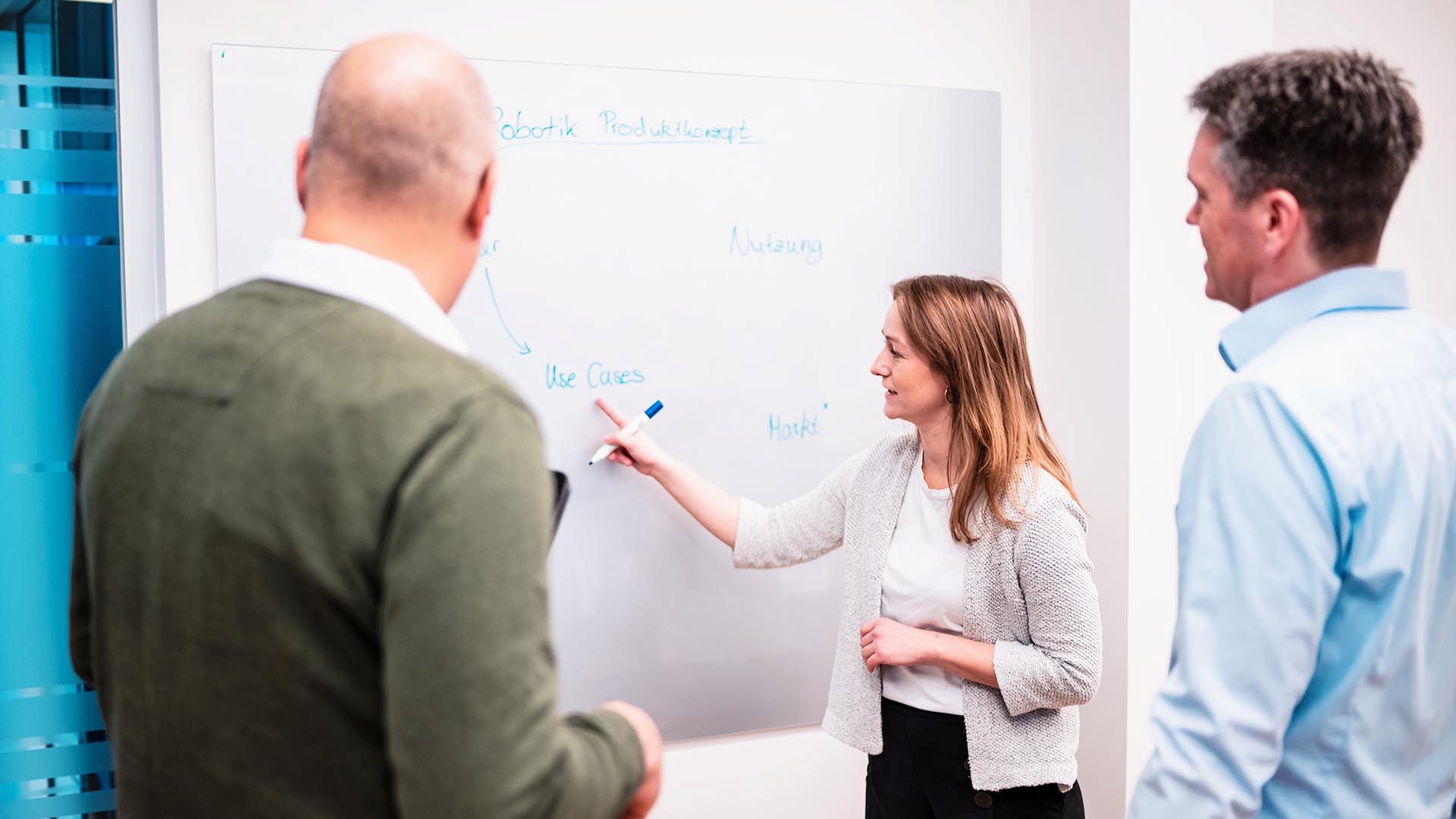 Eine Frau zeigt auf einem Whiteboard mit der Überschrift „Robotik Produktkonzept“ auf "Use Cases" und bespricht diese mit zwei Männern.