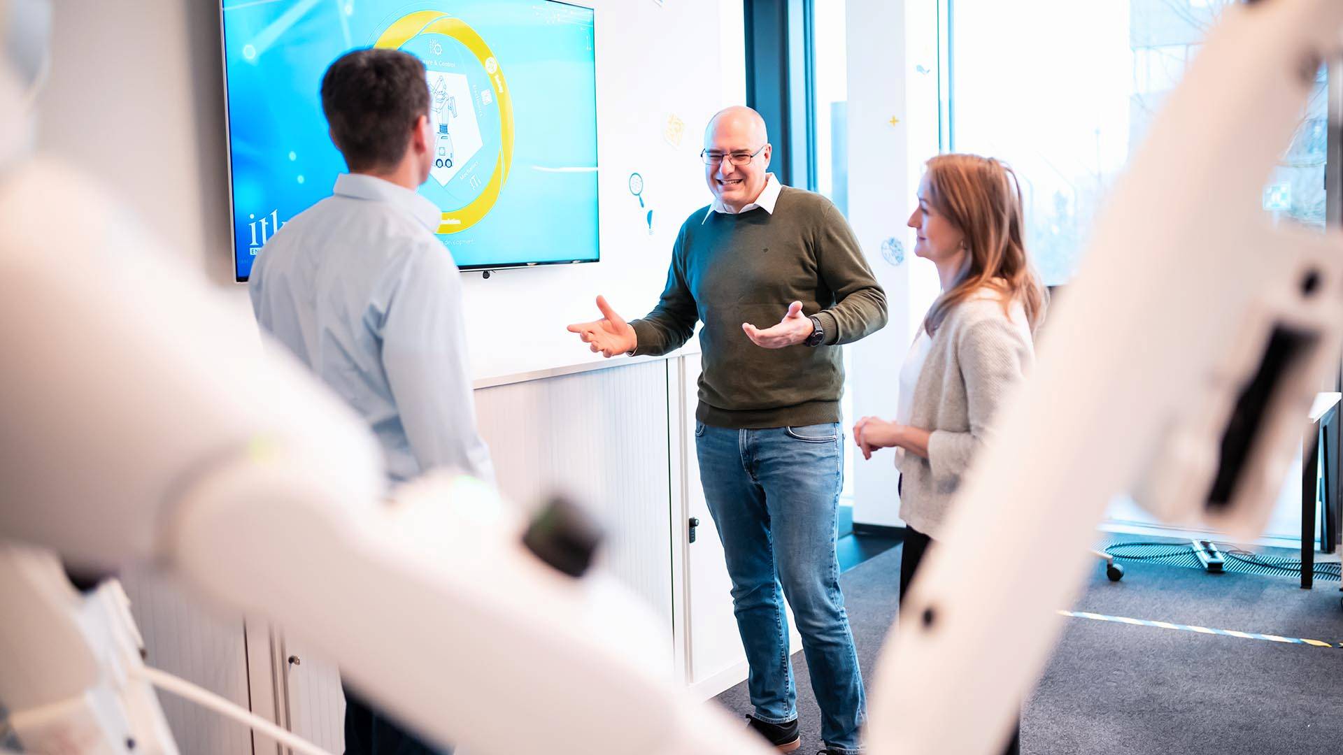 Drei Personen diskutieren vor einem Bildschirm, der "Robotics Consulting" anzeigt, und einem Roboter im Vordergrund.