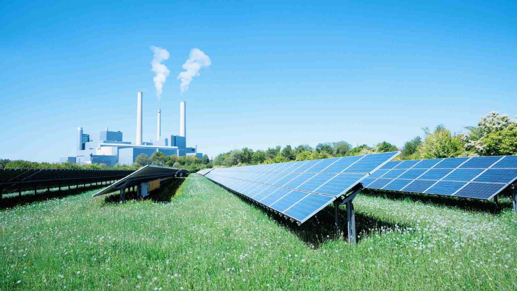 Solar panels in a field with smoking chimneys in the background.