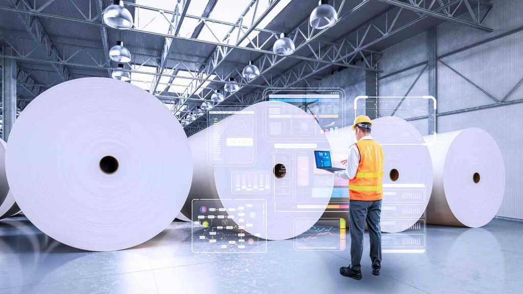 A man wearing an orange vest stands in front of large rolls of paper.